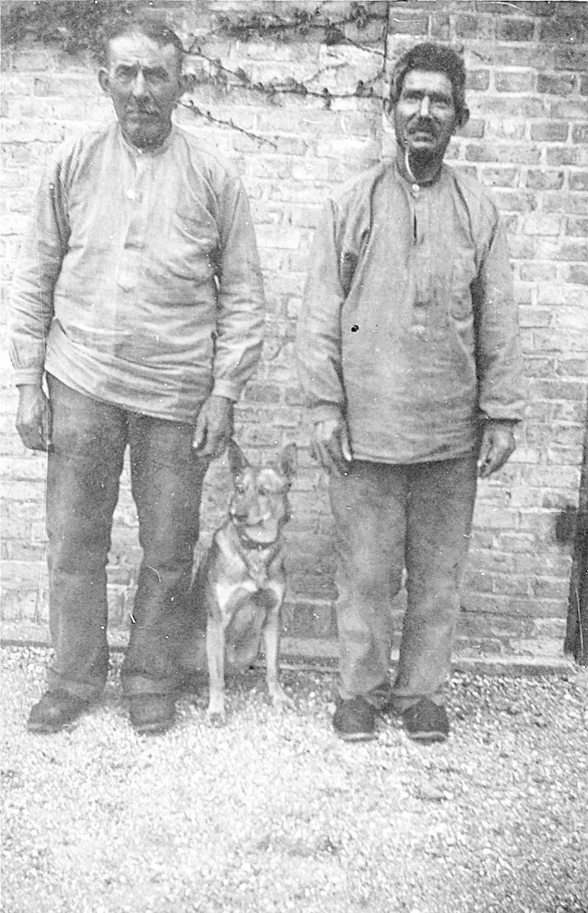 Fotografi af Kristen Andersen og Hans Jørgensen - taget i Fattiggårdens arbejdsafdeling i begyndelsen af 1940'erne. Hunden i midten er en af forvalterens politihunde, som han opdrættede på Fattiggården. Mændene er iklædt det typiske arbejdstøj til dens tids ”aktivering”: skærveknusning, brændehugning eller arbejde i et af værkstederne med for eksempel vævning af måtter til videresalg. - 