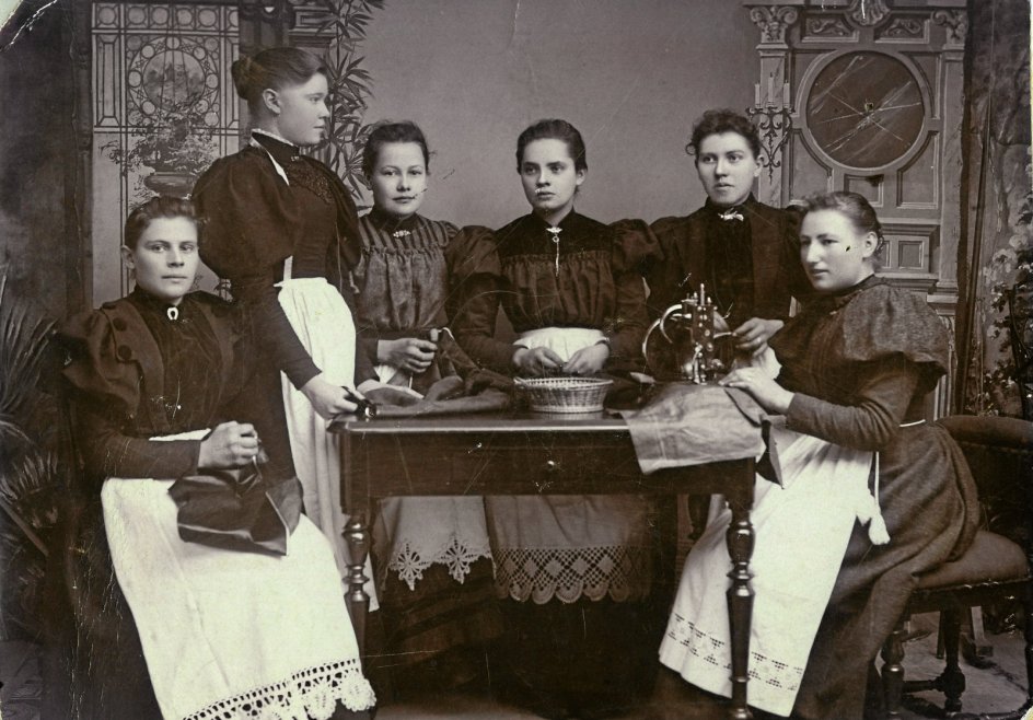 Omkring 1900 drev Kristiane Madsen med succes en systue i sit hjem i Storegade 26 i Grenaa. Her er Kristiane Madsen (stående) fotograferet sammen med sine ansatte i fotografens atelier, alle klædt i den nyeste mode – kjoler med skinkeærmer – og nystivede forklæder. Fotografiet var dermed en fin reklame for systuens tekniske formåen. Bemærk symaskinen til højre på fotoet. – Foto: Grenaa Egnsarkiv.